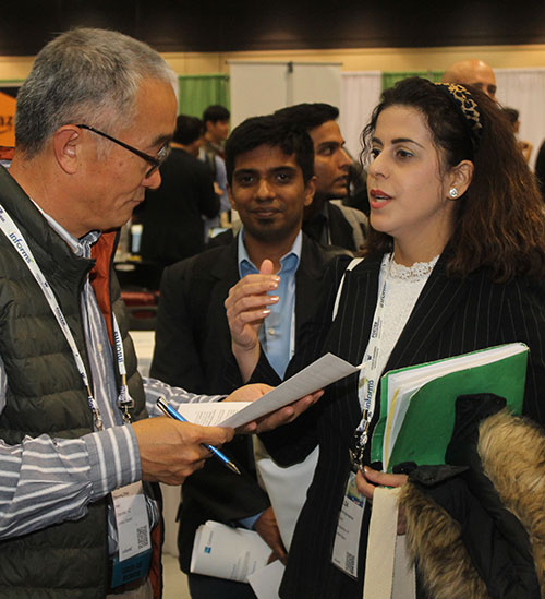 Two 2019 INFORMS Annual Meeting attendees, one an older asian man, the other a non-white woman, are in conversation over a paper. Behind them, a younger Indian man looks into the camera, smiling.